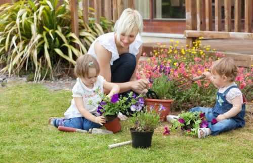 In this outdoor garden scene, a mature woman with short blonde hair, dressed in light clothing, is sitting on the well-maintained green lawn, engaging in planting and gardening activities alongside two young children. The children, a girl and a boy, are dressed casually; the girl in a white top and blue jeans, and the boy in a blue shirt and patterned trousers. They are working with pots containing a variety of flowering plants, including vibrant purple, pink, and yellow blooms, which they are either planting or arranging. The garden features a lush border of ornamental grasses and shrubs at the back, with a wooden deck or patio area visible nearby. The environment appears bright and clear, indicating a sunny day, enhancing the natural tones of the grass, plants, and wood surfaces. This scene exemplifies a family-friendly gardening activity in a typical residential garden, reflecting the outdoor maintenance and planting services provided by Garden Maintenance Palmers Green, situated in the N13 postal district. The setup emphasizes careful plant organization, gardening tools, and a tidy, inviting outdoor space suitable for leisure and family activities.