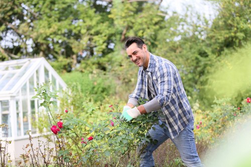 A man gardening in a lush backyard garden in Palmers Green, surrounded by vibrant pink and red flowering bushes, with a greenhouse visible on the left. The garden features a well-maintained lawn in the foreground, bordered by various shrubs and plants. Behind him, there are mature trees and dense greenery, providing a natural backdrop under soft daylight on a mild, partly cloudy day. The man is smiling as he tends to the plants, wearing casual clothing with gardening gloves, exemplifying professional outdoor maintenance suitable for services offered by Garden Maintenance Palmers Green. The scene captures a peaceful outdoor setting with natural tones of green, pink, and brown, emphasizing the importance of detailed garden care and landscaping services in the local area, including pruning, planting, and lawn maintenance.