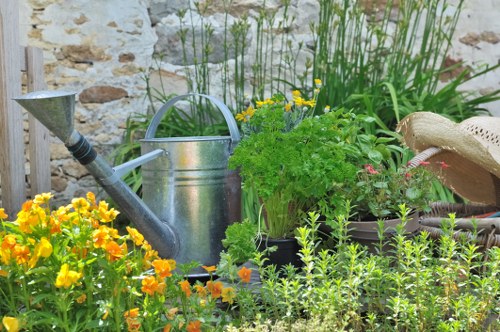 The image depicts a vibrant garden scene with a variety of plants and garden elements arranged in a small outdoor space. In the foreground, there are lush, dense flower beds bursting with bright yellow and orange blooms, adding color and visual interest. Behind the flowers, a weathered stone wall provides a rustic backdrop, complementing the natural setting. A metal watering can sits near the plants, suggesting ongoing garden maintenance, while a grey plastic or metal container is partly visible among the greenery. Tall, leafy green plants, possibly ornamental grasses or herbs, grow towards the top of the scene, creating vertical accents. To the right, part of a woven sun hat rests on a surface, indicating a gardener taking a break or preparing for work. The environment is well-lit, suggesting a sunny day, with natural light illuminating the various textures and shades of green, yellow, and earth tones. Overall, this scene exemplifies a well-maintained, lively garden space suitable for services like lawn care, planting, and outdoor maintenance for the Palmers Green area, with subtle ties to local gardening practices and styles.