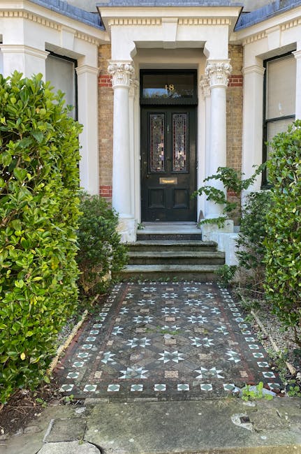 A front garden leading to a black door with a gold mail slot, framed by ornate white columns supporting a structured overhang. The pathway to the entrance features decorative patterned tiles with a ge